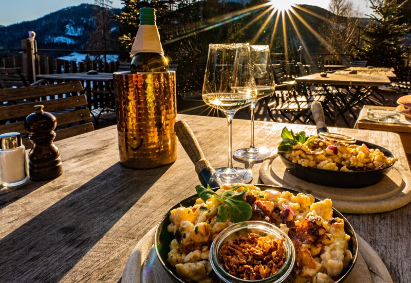 Outdoor dining setup with wine and gourmet dishes on a sunlit terrace overlooking wintery mountains at sunset.