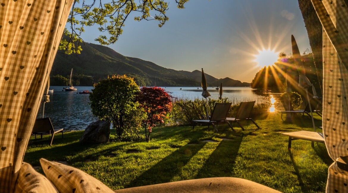 Blick auf den Fuschlsee bei Sonnenuntergang, mit Liegestühlen auf einer Wiese, Segelbooten im Wasser und Bergen im Hintergrund.