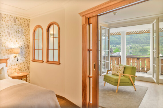 Hotel room with a cozy armchair and balcony offering a scenic view of lush, green hills.