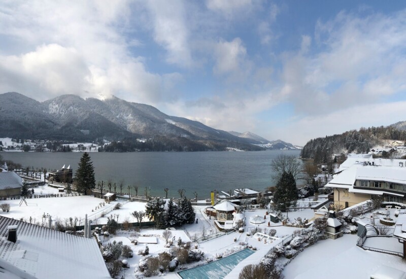 Snow-covered landscape with a lake and mountains in the background, surrounded by buildings and trees under a partly cloudy sky.