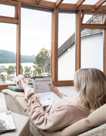 Two people in robes relax in a spa lounge with panoramic lake and mountain views.