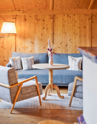 Cozy wooden living area with a blue sofa, armchairs, and a round table adorned with a vase of dried flowers, softly lit by a lamp.