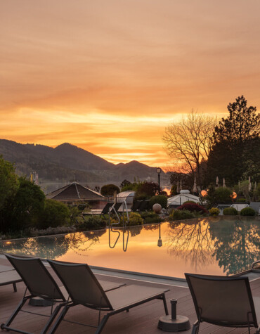 Sunset view over a tranquil pool with mountains in the background at Waldhof Fuschlsee Resort, featuring lined-up lounge chairs.