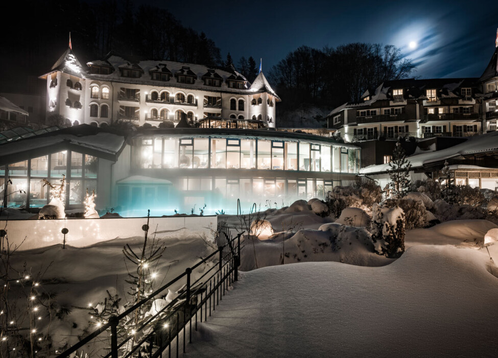 Snow-covered hotel at night, warmly lit with decorative lights and a glowing pool, under a full moon.