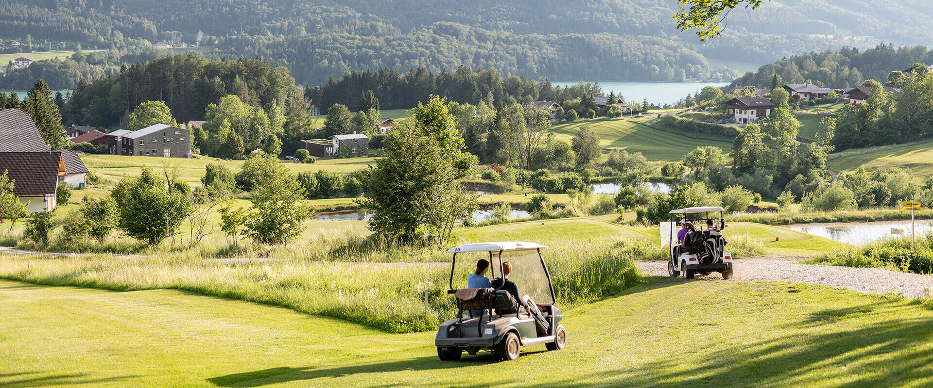 Golf carts on a lush, scenic course with mountains and a lake in the background.