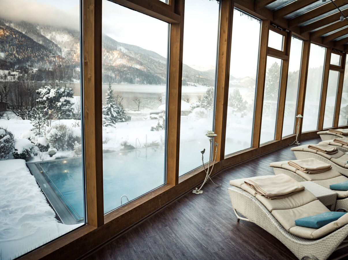 Lounge chairs by large windows overlooking a snow-covered scenic landscape with a pool and mountains in the background.