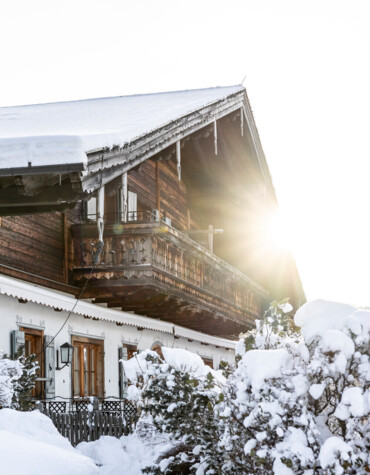 A snow-covered chalet with a wooden balcony, surrounded by snowy trees. The sun peeks through, illuminating the winter landscape.