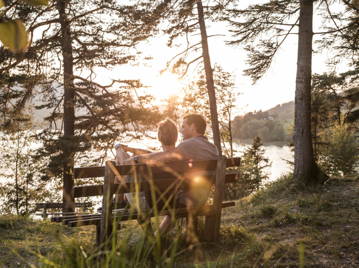 A couple sits on a bench in a forest, overlooking lake Fuschl, surrounded by trees.