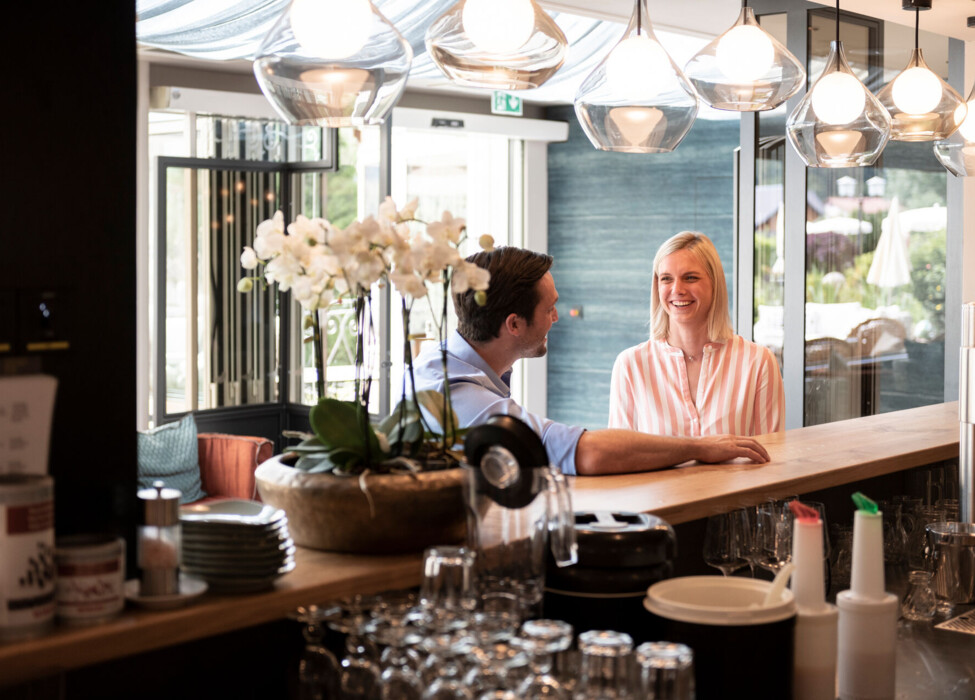A man and woman smiling at each other in a cozy café, with elegant lighting and a potted plant on the counter.