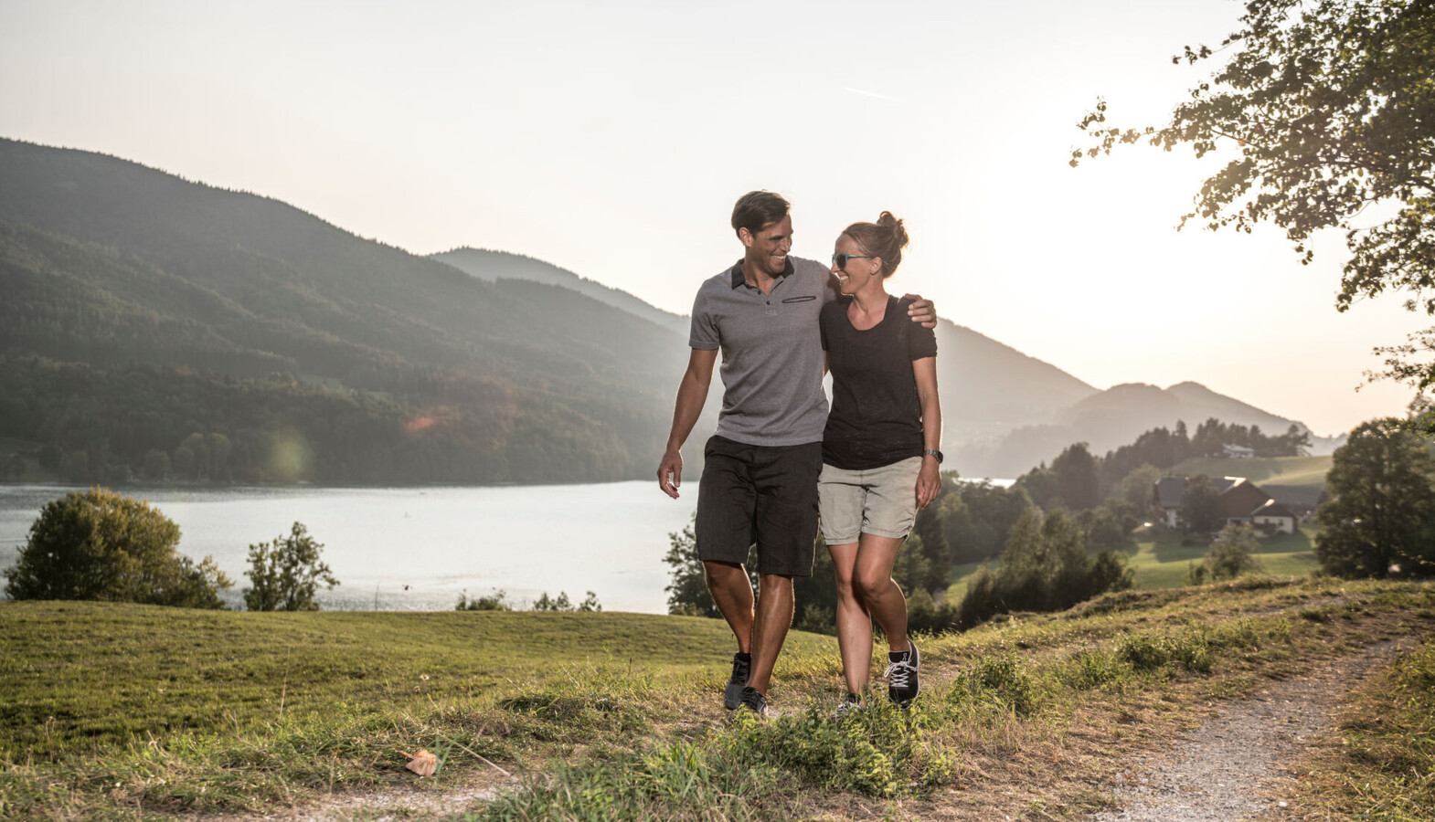 A couple walks along a trail next to lake Fuschl at sunset, with mountains in the background near Waldhof Fuschlsee Resort.